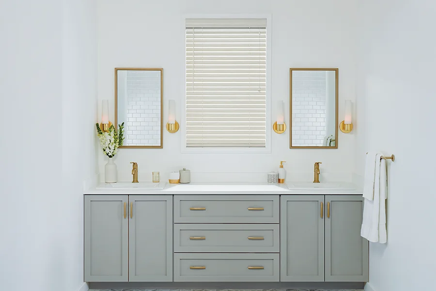 Durable, water-resistant faux-wood blinds are perfect for bathrooms. White faux-wood window blind in a white bathroom with a grey double vanity